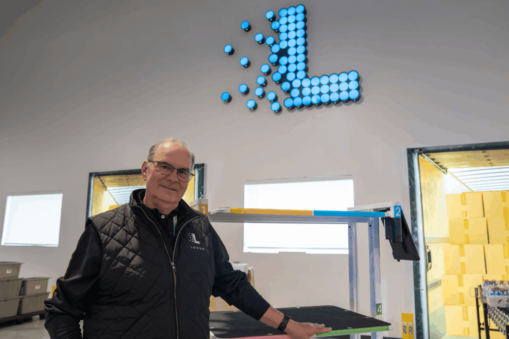 Locus Robotics team member in a branded vest beneath an illuminated Locus logo, hand resting on a picking station