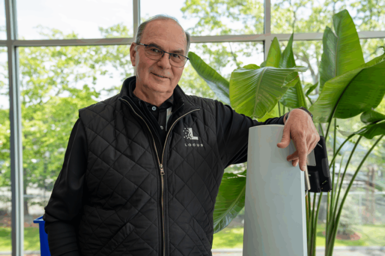 Locus Robotics team member in a branded quilted vest leaning on a LocusBot mast in front of large tropical plants