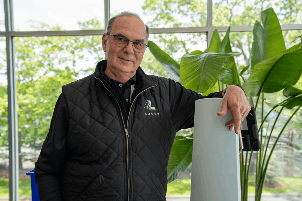 Locus Robotics team member in a branded quilted vest leaning on a LocusBot mast in front of large tropical plants
