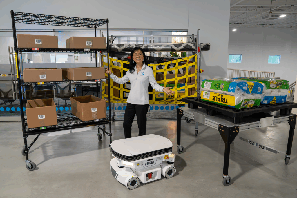 Locus Robotics employee gesturing beside a low-profile autonomous robot, shelving cart and pallets of paper goods