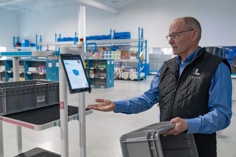 Locus Robotics team member in a branded vest holding a gray tote and gesturing to a robot's touchscreen display