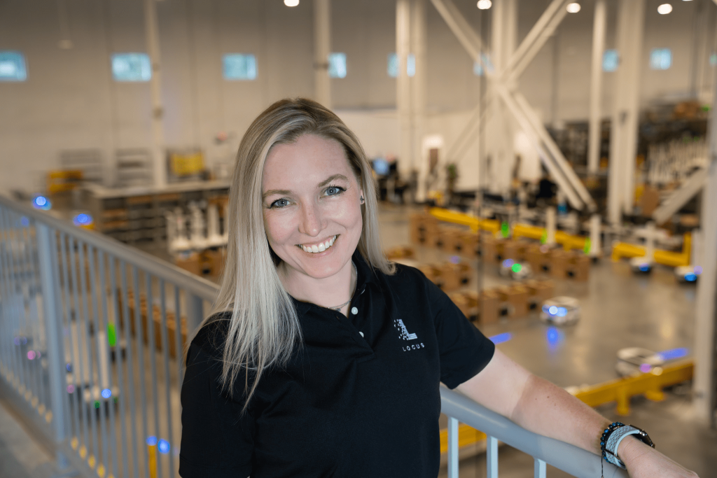 Smiling Locus Robotics employee on a mezzanine overlooking a warehouse floor with LocusBots and conveyors