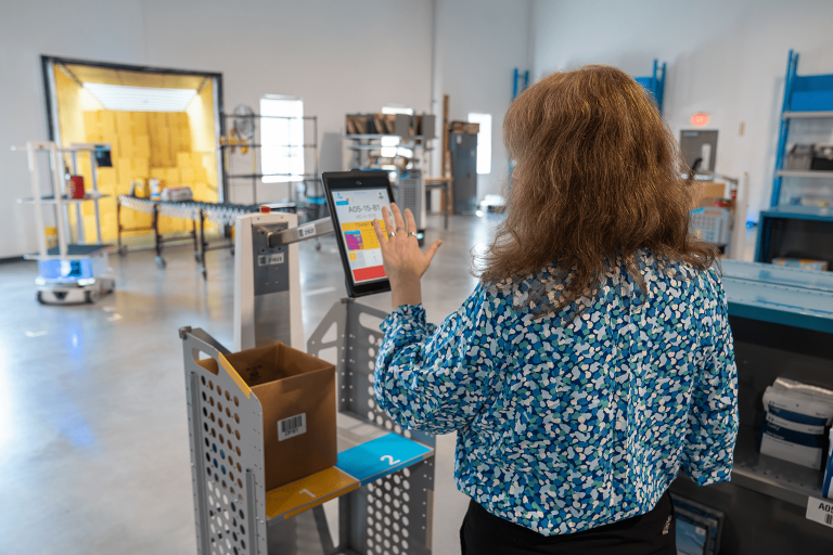 Warehouse worker tapping a LocusBot touchscreen showing pick instructions beside a cart holding a cardboard box
