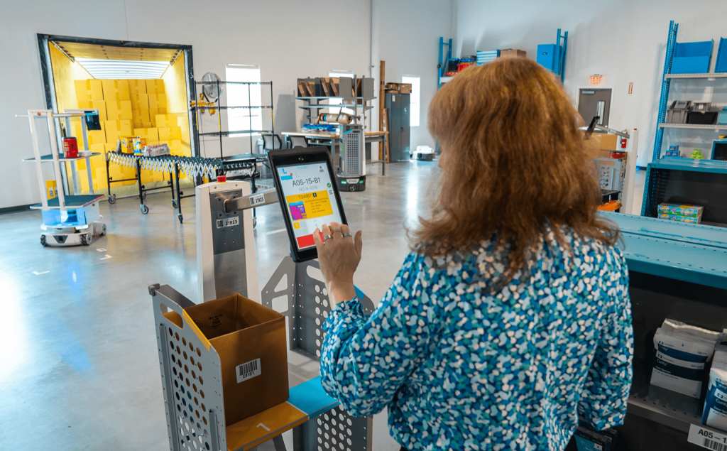 Worker scanning a tablet on a Locus robot to pick an item into a box in a bright warehouse