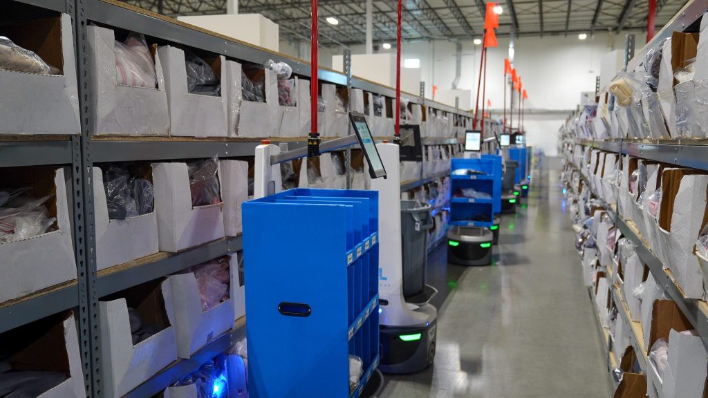 Row of LocusBots carrying blue totes along shelving aisles filled with bagged apparel in an order-picking area