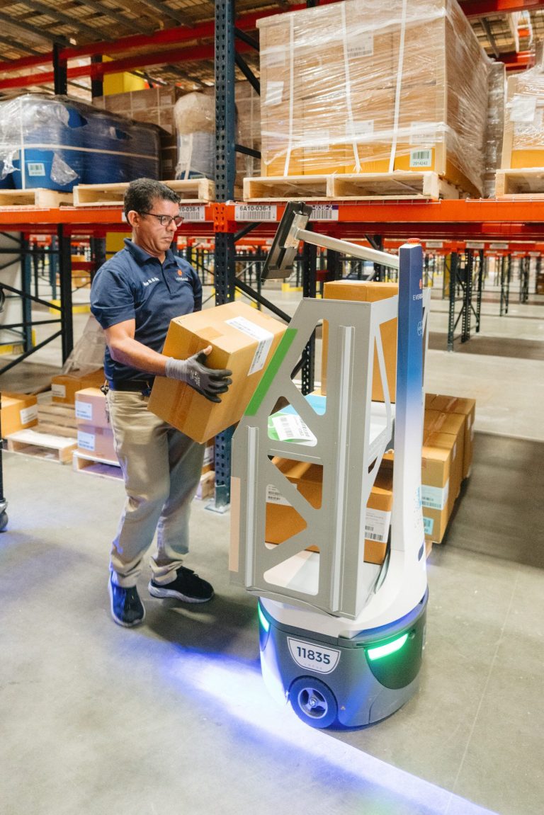 Warehouse worker lifting a cardboard box onto a Eversana-branded LocusBot beside pallet racking