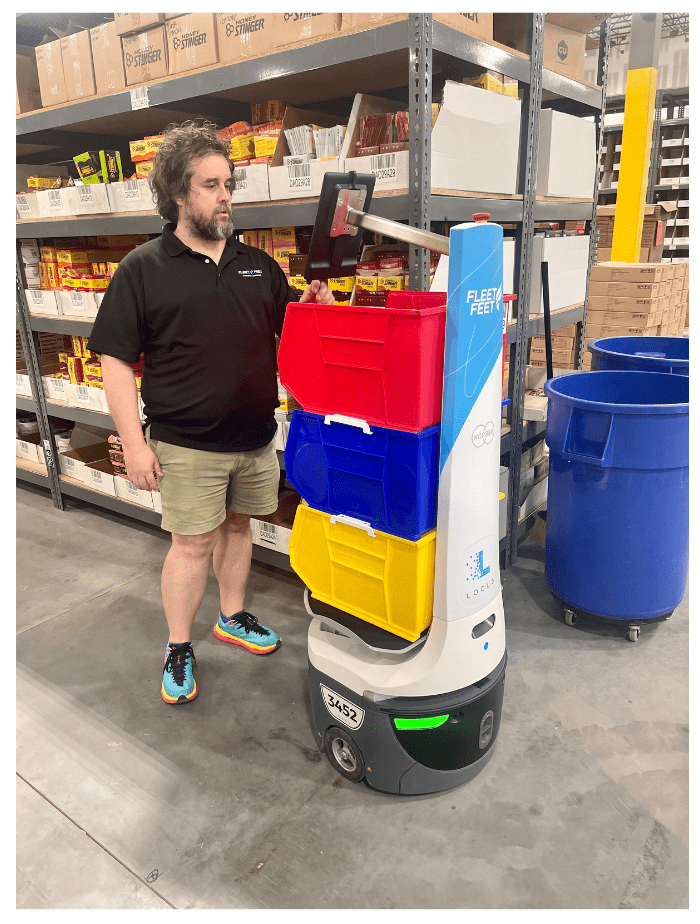 Fleet Feet associate beside a LocusBot carrying red, blue and yellow totes in a warehouse aisle