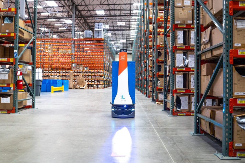 LocusBot with an orange and white mast driving down a long warehouse aisle lined with stocked pallet racking