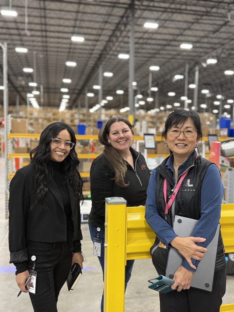 Three smiling professionals standing by a yellow guardrail in a large, brightly lit fulfillment warehouse