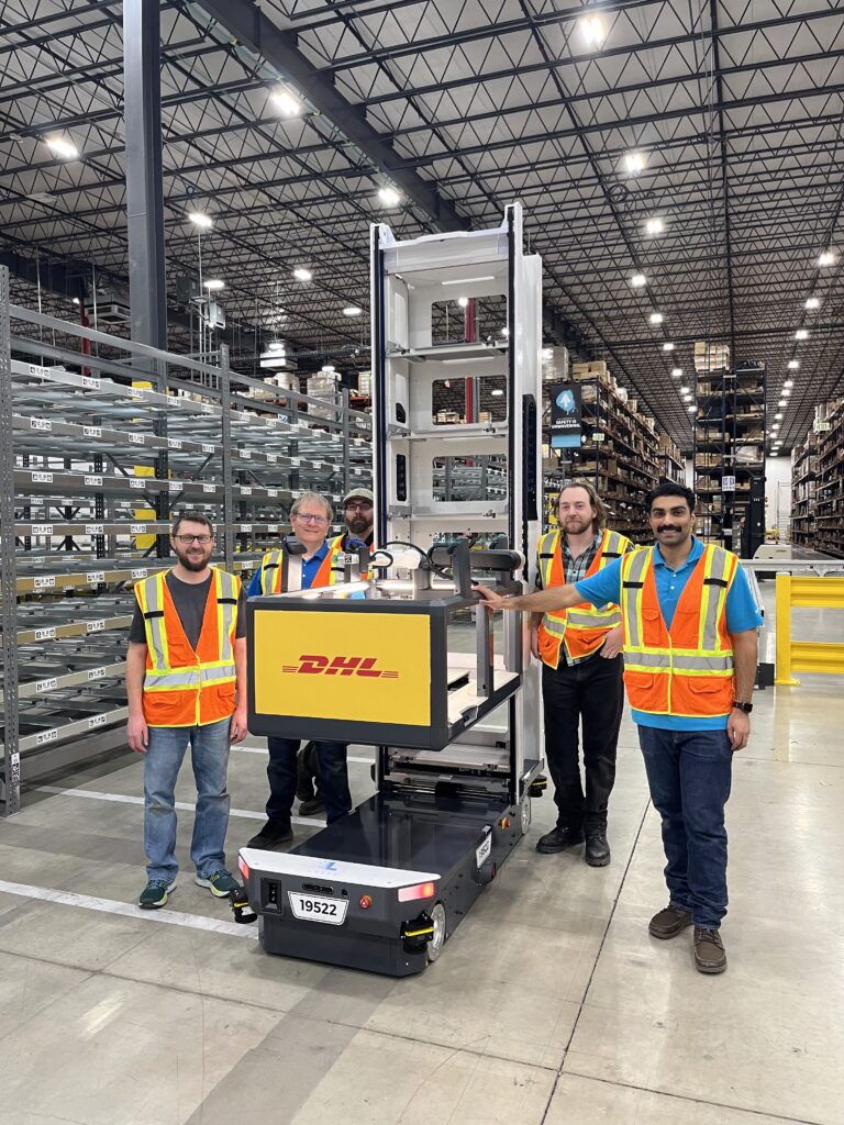 Five team members in safety vests standing with a DHL-branded Locus Array robot in a warehouse aisle