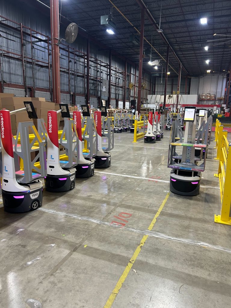 Row of Staples-branded LocusBots parked in a warehouse beside yellow safety barriers