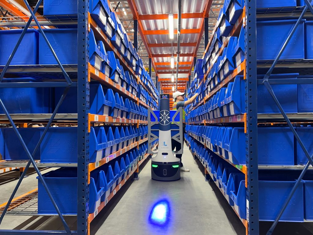 LocusBot in an aisle of blue storage bins as a worker in a cap reaches up to a shelf