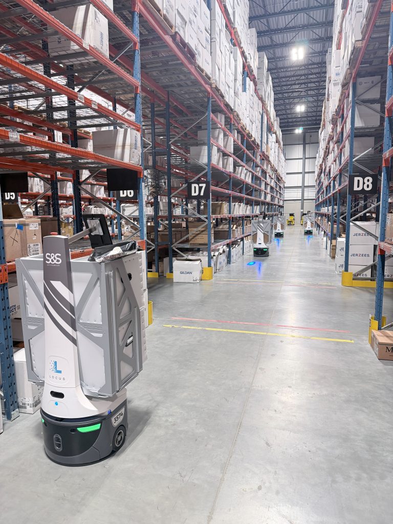 S&S Activewear-branded LocusBots moving through tall warehouse aisles of racking stocked with apparel boxes
