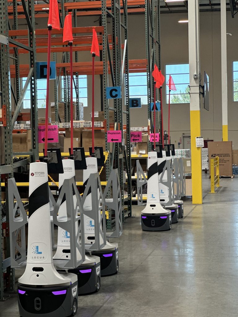 Row of Motivational Fulfillment-branded LocusBots with orange flags near Pick and Replenish signs in a warehouse