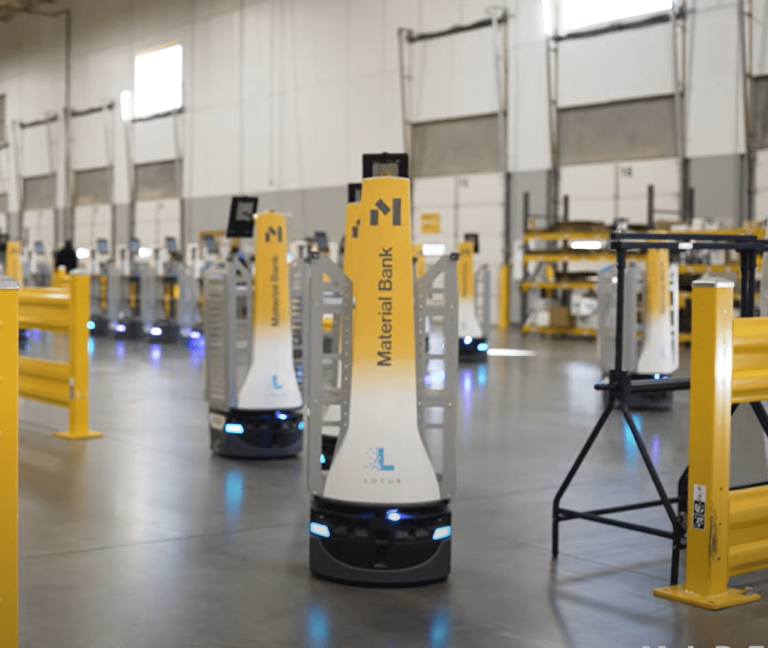 Yellow and white Material Bank-branded LocusBots moving across a warehouse floor behind yellow safety railings