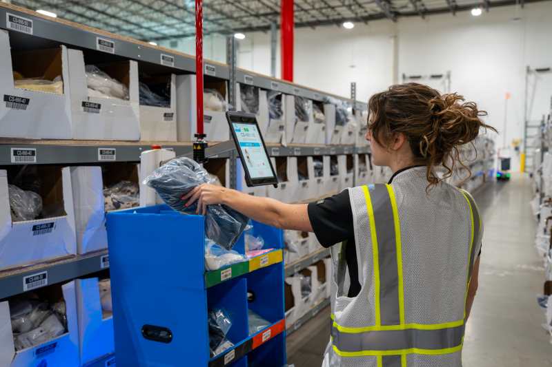 Warehouse picker in a safety vest placing a bagged item into a blue cart on a Locus Origin robot