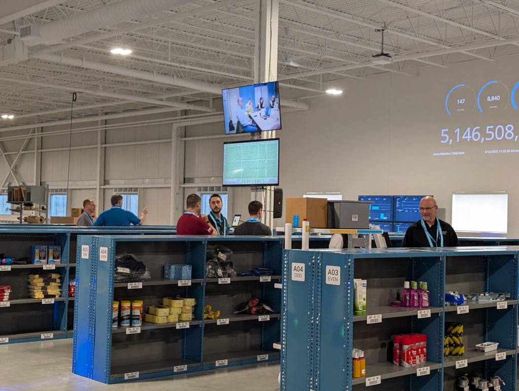 Visitors observing a live warehouse demo with blue pick shelving and a large live throughput dashboard
