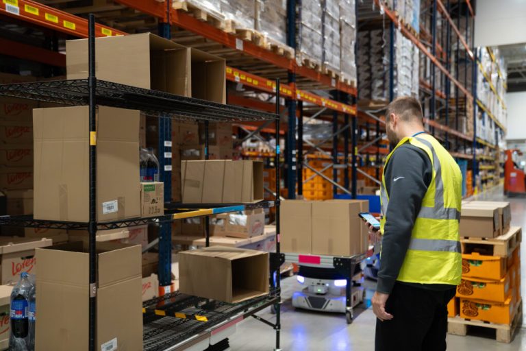 Worker in a safety vest using a handheld device beside a Locus Vector robot carrying a loaded shelving cart