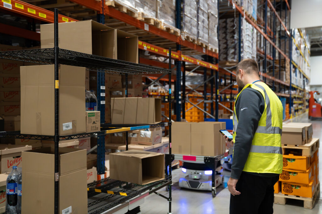 Worker in a safety vest using a handheld device beside a Locus Vector robot carrying a loaded shelving cart