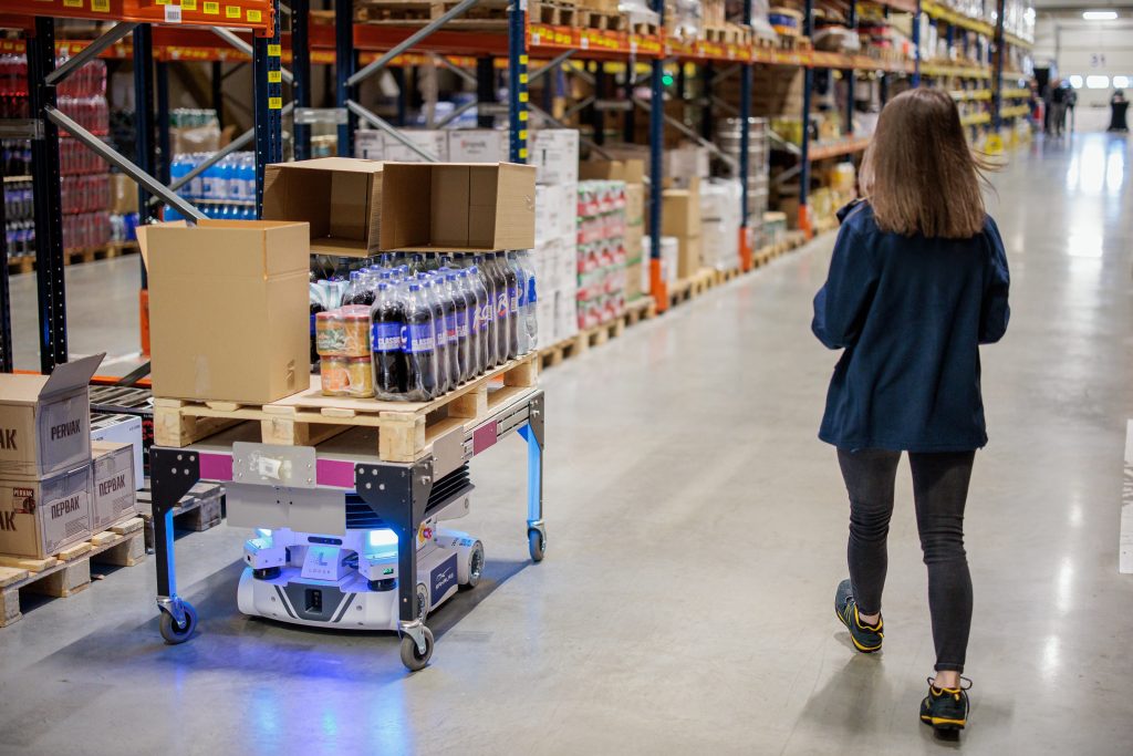 Warehouse worker walking ahead of a Locus Vector robot carrying a pallet of bottled drinks and boxes