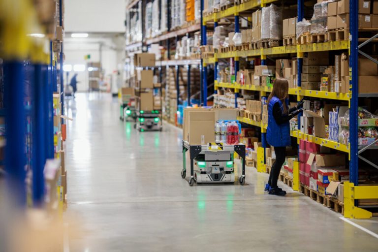 Worker picking from grocery shelving beside Locus Vector robots carrying carts of boxes down an aisle