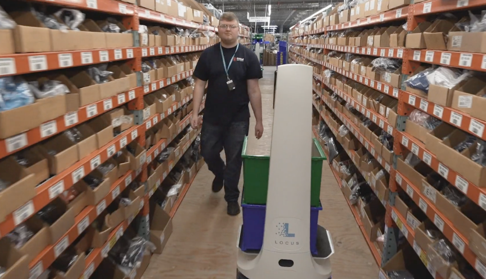 Associate walking behind a LocusBot carrying green and blue totes down a narrow aisle of bin shelving