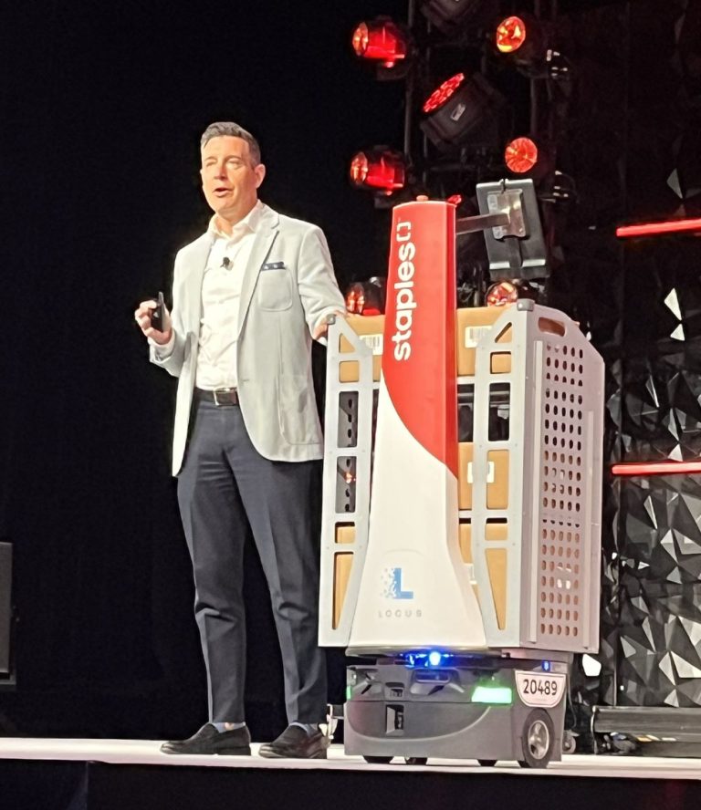 Speaker presenting on a stage beside a Staples-branded LocusBot loaded with cardboard boxes