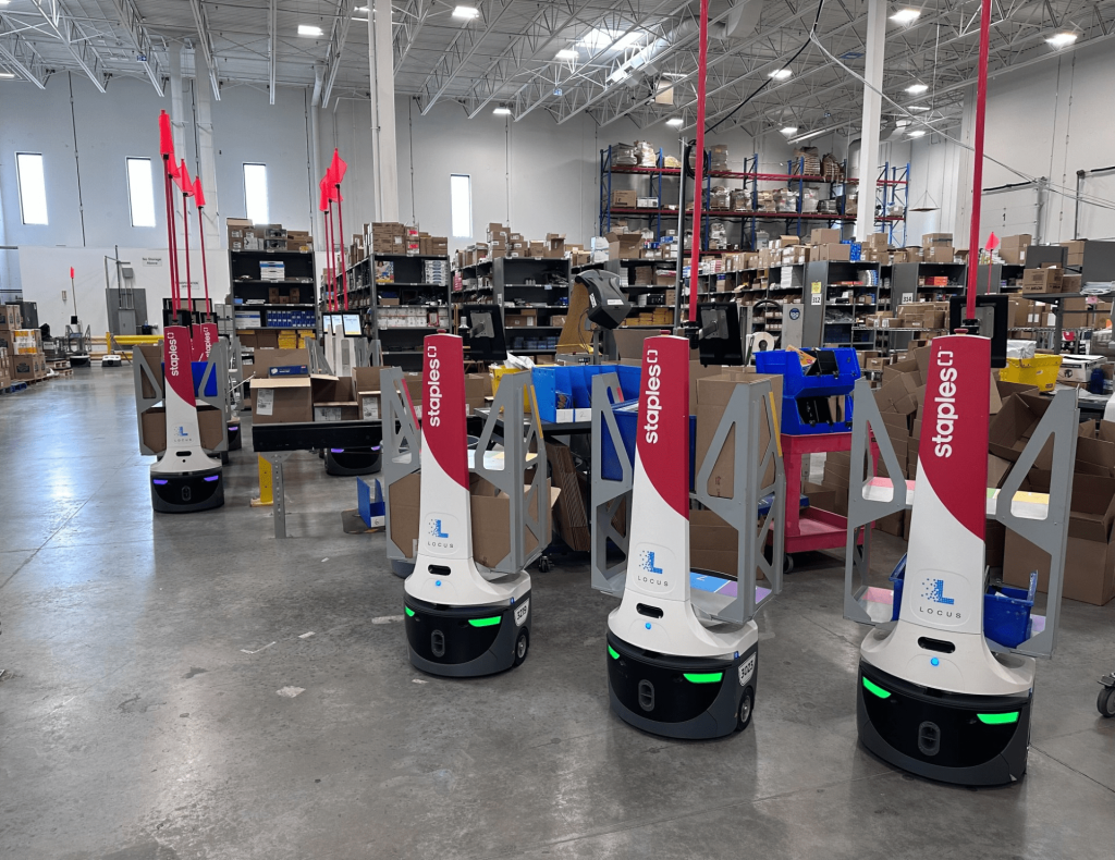 Staples-branded LocusBots waiting among shelving and packing stations in a busy fulfillment center