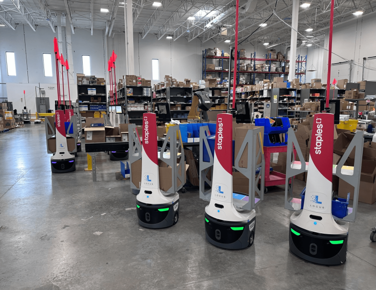 Staples-branded LocusBots waiting among shelving and packing stations in a busy fulfillment center