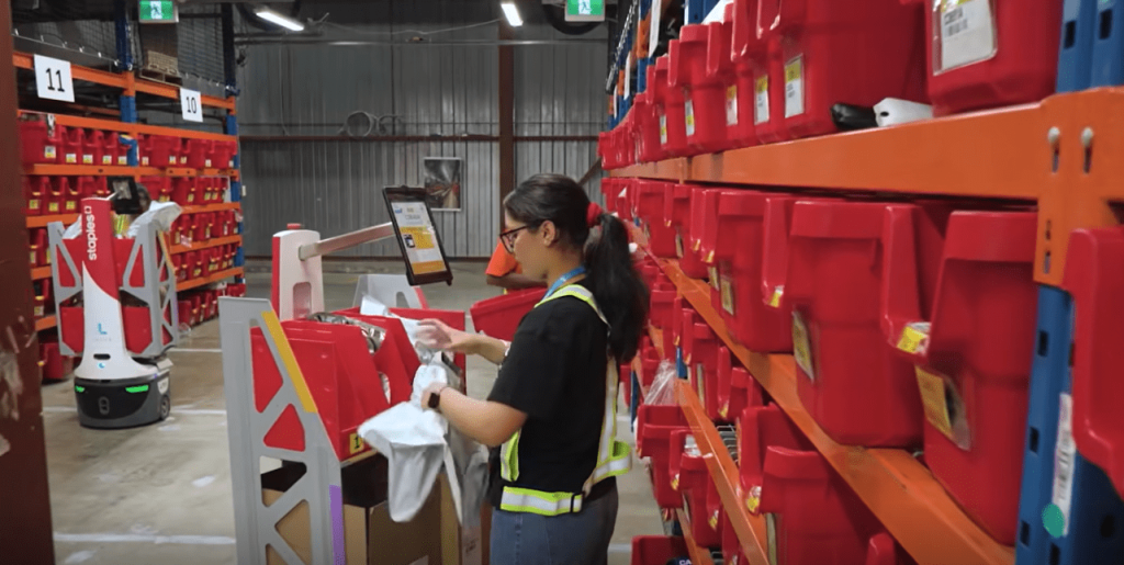 Associate in safety vest picking an item into a tote on a Staples-branded LocusBot in a red-bin aisle