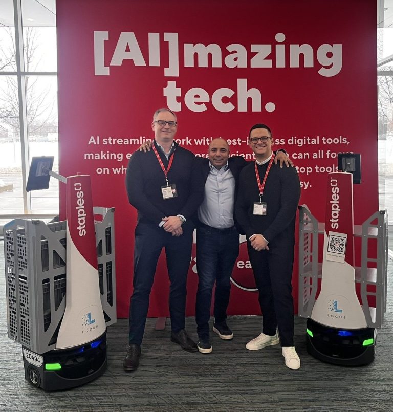 Three Staples Canada team members posing between two Staples-branded LocusBots at an [AI]mazing tech display