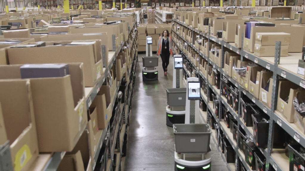 Associate walking an aisle of cardboard bin shelving with several LocusBots carrying gray totes