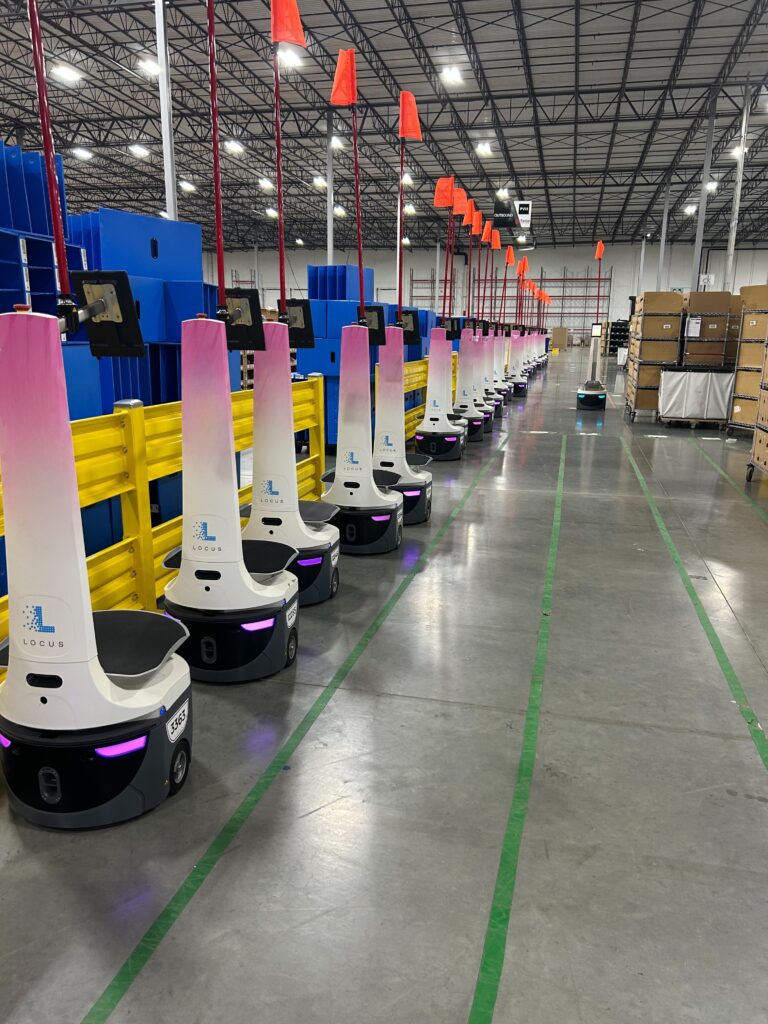 Long line of pink and white Radial-branded LocusBots with orange safety flags lined up in a warehouse