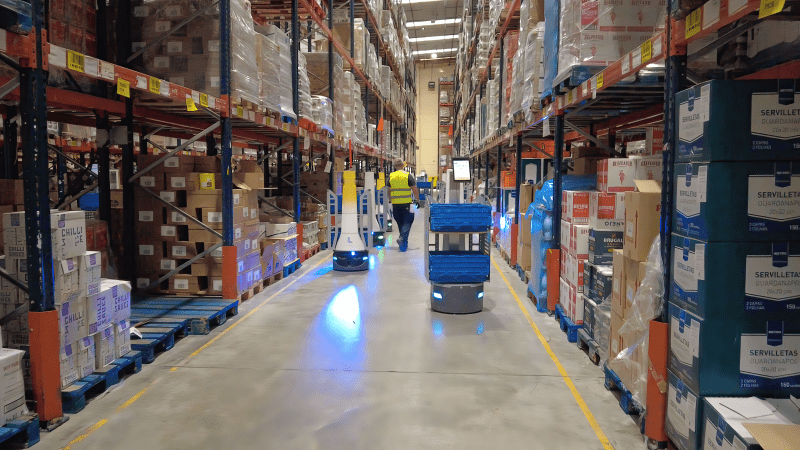 LocusBots and a worker in a high-visibility vest moving down a long aisle of stocked pallet racking