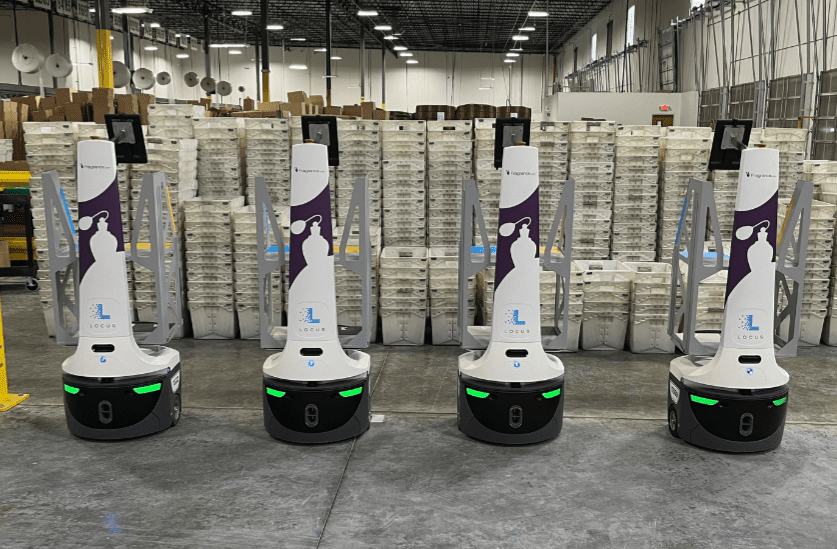 Four LocusBots with fragrance bottle graphics lined up in front of stacked totes in a warehouse