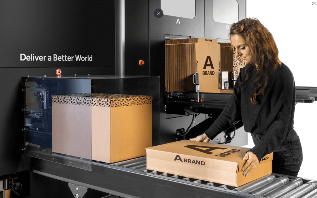 Worker placing a branded shipping box on a conveyor at an automated box-making packaging machine