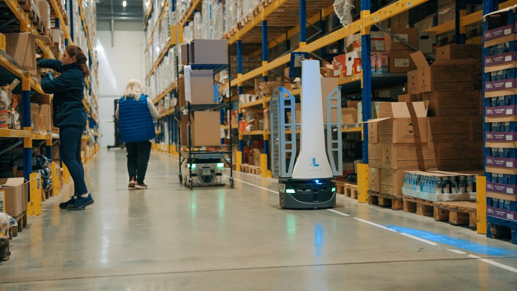 Associates picking in a Sakalas food warehouse aisle alongside a Locus Origin robot and a Locus Vector