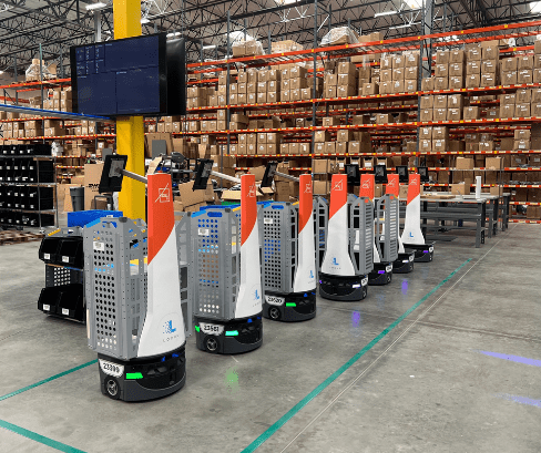 Row of LocusBots with orange masts and gray totes lined up in front of warehouse racking